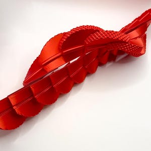 Pu&ograve; includere: Una scultura astratta rosso vivo con un design a nastro fluente. Il pezzo presenta una sezione strutturata, simile a un tessuto, e diversi elementi curvi a strati. La scultura &egrave; ambientata su uno sfondo bianco, evidenziando i suoi dettagli e la sua forma.