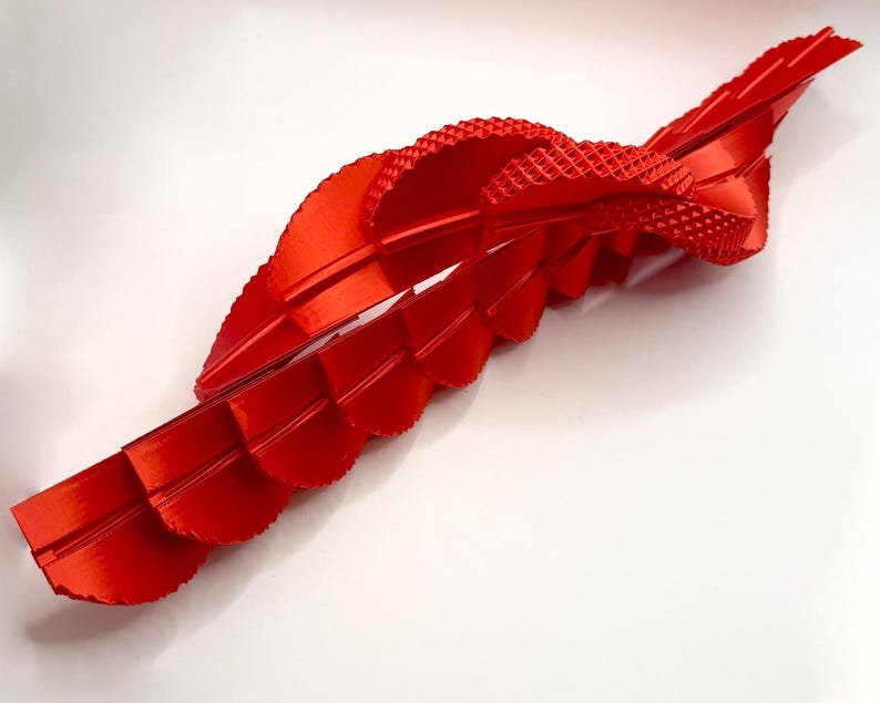 Pu&ograve; includere: Una scultura astratta rosso vivo con un design a nastro fluente. Il pezzo presenta una sezione strutturata, simile a un tessuto, e diversi elementi curvi a strati. La scultura &egrave; ambientata su uno sfondo bianco, evidenziando i suoi dettagli e la sua forma.