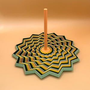 Könnte beinhalten: Eine geometrische, abstrakte Skulptur mit einem zentralen orangefarbenen Pfosten. Die Basis ist ein mehrschichtiges, sternförmiges Design mit abwechselnden Bändern in Gelb, Schwarz und Grün. Der Hintergrund ist ein helles Orange.