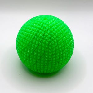 Op de afbeelding: Een heldere neongroene bol met een getextureerd oppervlak dat bestaat uit een raster van kleine, verhoogde vierkante bultjes. Het object heeft een duidelijke verticale naad die van de bovenkant naar het midden loopt en een geometrisch patroon vormt.