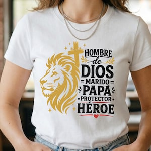 May include: A white t-shirt featuring a gold lion head graphic, a cross, and black text that reads HOMBRE de DIOS MARIDO PAPA PROTECTOR HEROE. The design includes small gold stars, decorative leaf accents, and a small red heart at the bottom.