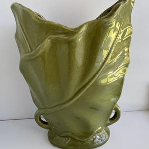 May include: A glossy olive green ceramic vase shaped like a large, folded leaf. The vessel features a wide, undulating rim, a textured surface with prominent veins, and two small, circular handles positioned near the base on either side.