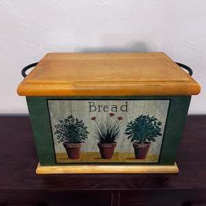 May include: A rectangular wooden bread box with a natural wood finish lid and base. The front panel is painted dark green and features a graphic of three potted herbs with the text "Bread" above them and "Parsley", "Chives", and "Mint" below.