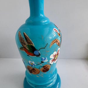 May include: A turquoise-colored decorative vase with a flared base and a slender neck. The vase is adorned with hand-painted details, including a colorful bird, flowers, and foliage. The design features shades of red, brown, white, and blue.