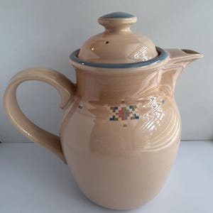 May include: A beige ceramic teapot with a matching lid, featuring a blue rim and a small, multi-colored geometric pattern on the front. The teapot has a curved handle, a short spout, and a smooth, glossy finish throughout its entire surface.