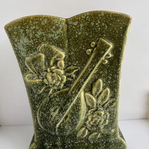 May include: A ceramic vase with a mottled olive green glaze featuring a raised relief design of a stringed musical instrument, likely a mandolin or lute, surrounded by detailed floral motifs including blooming roses and leaves on the front surface.