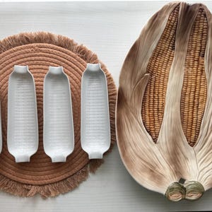 May include: Four white ceramic corn-shaped dishes arranged on a round tan woven placemat with fringe, alongside a large oval ceramic serving platter shaped like two ears of corn with realistic brown and yellow kernels and tan husks.