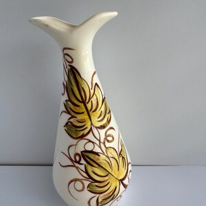 May include: A white ceramic vase with a flared, asymmetrical rim and a tapered body. The surface features hand-painted yellow and green leaves with brown outlines and swirling vine details extending vertically along the front of the piece.