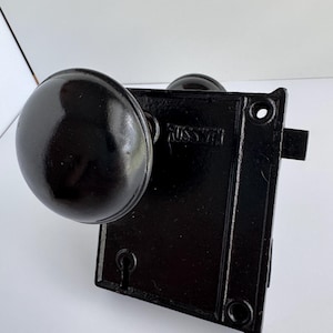 May include: A black door knob and lock set with a round knob and rectangular backplate. The lock set has the word "RUSSWIN" embossed on the backplate. The hardware is made of metal and has a glossy finish.