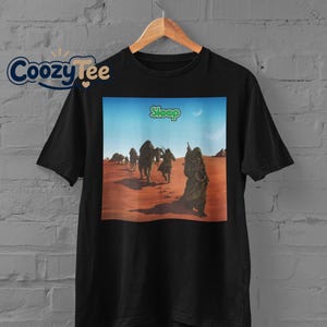 Peut inclure: Un t-shirt noir à manches courtes avec une impression graphique représentant un paysage désertique avec des personnages et des chameaux sous un ciel bleu. Le mot "Sleep" est imprimé en lettres vertes au-dessus. Le t-shirt est sur un cintre.