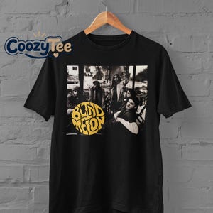 Peut inclure: Un t-shirt noir à manches courtes avec une impression graphique du groupe Blind Melon. Le motif comprend une photographie en noir et blanc des membres du groupe avec un logo circulaire jaune proéminent portant le texte Blind Melon.