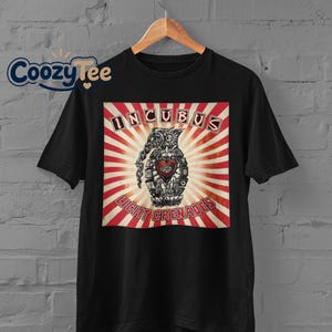 May include: A black short-sleeved t-shirt featuring a graphic print of a hand grenade with a heart and eye symbol. The design includes the text INCUBUS and LIGHT GRENADES set against a background of red and white radiating sunburst stripes.