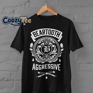Puede incluir: Camiseta negra con un gráfico blanco. El gráfico presenta la palabra "BEARTOOTH" en una pancarta, un diseño circular con la letra "B" y la palabra "AGGRESSIVE" debajo. También se incluye un par de cuchillos cruzados.