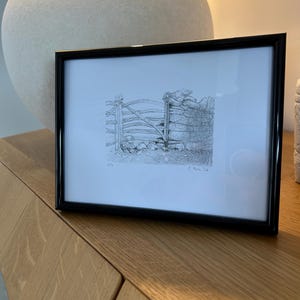 Puede incluir: Un boceto en blanco y negro enmarcado de una puerta de madera rústica y un muro de piedra. La obra de arte está en un marco negro y descansa sobre una superficie de madera clara. Un objeto grande, esférico y texturizado está en el fondo.
