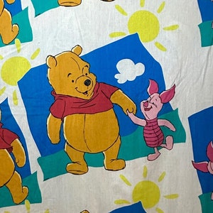 May include: White fabric with a repeating pattern of Winnie the Pooh and Piglet. Winnie the Pooh is wearing a red shirt and Piglet is wearing a pink shirt. The background is blue with white clouds and yellow suns.