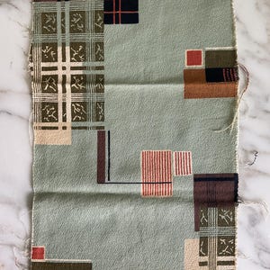 May include: A rectangular piece of sage green fabric featuring a geometric pattern of scattered squares and rectangles in shades of brown, black, cream, and rust orange, with some sections displaying a plaid design and others showing fine line details.
