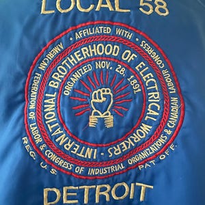 May include: A blue jacket featuring an embroidered emblem on the back. The text reads LOCAL 58, DETROIT, and INTERNATIONAL BROTHERHOOD OF ELECTRICAL WORKERS, ORGANIZED NOV. 28, 1891, with a central fist graphic and red and gold circular border details.
