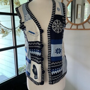 May include: A sleeveless knit vest featuring a winter theme with a color palette of navy blue, light blue, and white. The vest displays various patterns including snowflakes, mittens, and sleds, accented with small white beads and dark blue buttons.