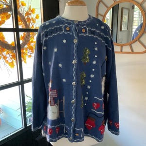 May include: A blue cardigan sweater with winter-themed embroidery. The sweater features a snowman, Christmas trees, snowflakes, and the words "Winter Snowman". It has heart-shaped buttons and red mitten and heart details.