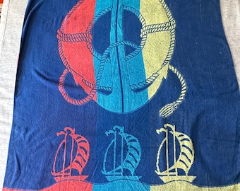May include: A navy blue beach towel featuring nautical designs. The towel has a large anchor with a red, blue, and yellow life preserver design. Below are three sailboats above a wavy stripe design in red, blue, and yellow.