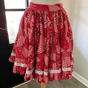 May include: A red and white knee-length skirt featuring a patchwork design of bandana prints and gingham patterns. The skirt has a gathered waistband and two horizontal tiers of white eyelet lace trim along the lower portion of the garment.