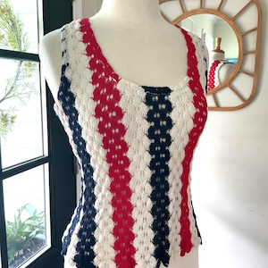 Puede incluir: Una camiseta de tirantes de ganchillo sin mangas con rayas verticales en colores alternos blanco, rojo y azul marino. La prenda tiene un escote redondo y un patrón de punto abierto y texturizado, exhibida sobre un torso de maniquí.