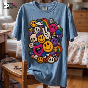 Könnte beinhalten: Ein hellblaues T-Shirt mit einem großen, zentralen Grafikdruck, der aus verschiedenen bunten Elementen besteht, darunter mehrere gelbe Smileys, ein weißes Friedenszeichen, Totenköpfe, ein rosa Herz, ein Regenbogen, Wolken und kleine Blumen.