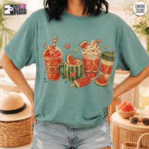 May include: A teal short-sleeve t-shirt featuring a graphic print of various watermelon-themed treats, including a smoothie, a mug, a whipped cream-topped drink, a coffee cup, and a popsicle, accented with small red and green heart shapes.