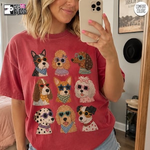 Puede incluir: Una camiseta roja de manga corta con un estampado gráfico de nueve razas de perros diferentes, cada una con gafas de sol coloridas y pañuelos o collares estampados. La camiseta tiene un aspecto lavado de estilo vintage.