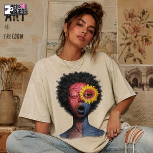May include: Beige t-shirt featuring a split-face graphic with a sunflower eye. The face is divided into red and blue halves, with black curly hair. The top left corner of the image has the text "MASTERPIECE ART IS FREEDOM".