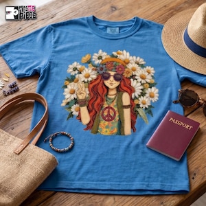 May include: A blue t-shirt featuring a graphic illustration of a person with long red hair, wearing sunglasses and a peace sign necklace, surrounded by white daisies. The shirt has a crew neckline and short sleeves, with the text Mastee Piece visible.