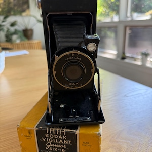 May include: A vintage black Kodak Vigilant Junior SIX-16 camera with a Kodet lens, resting on its original yellow box. The box features the Kodak logo and product details. The camera is open, revealing its lens and bellows.
