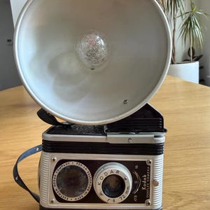 May include: Vintage Kodak Duaflex camera with a large, silver flash attachment. The camera body is black and silver, with the Kodak logo and text visible. The lens has f/8 and 72 mm markings. A black strap is attached to the side. Made in Rochester, NY, USA.