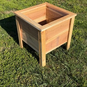 May include: A square wooden planter box with a natural finish. The planter has a raised design and is supported by four legs. The box is made of light brown wood and sits on green grass.