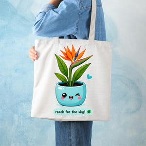Op de afbeelding: Een witte canvas draagtas met een afbeelding van een paradijsvogelbloem in een lichtblauwe pot met een knipogend gezicht. Het ontwerp bevat een klein blauw hartje en de tekst reach for the sky! met een klein groen blaadje onder de pot.