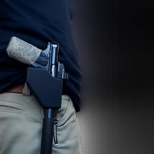 Pu&ograve; includere: Una pistola nera in una fondina nera &egrave; parzialmente visibile, infilata nella cintura di pantaloni color cachi. L'impugnatura della pistola &egrave; avvolta in un motivo mimetico. La persona indossa una camicia blu navy.