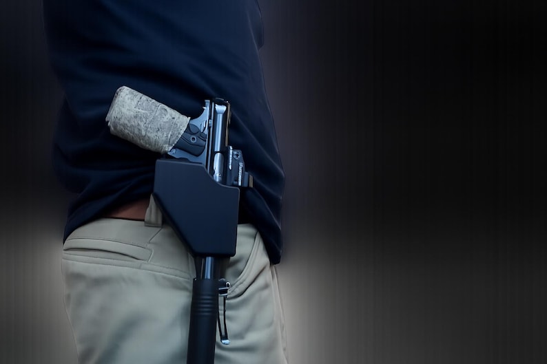 Pu&ograve; includere: Una pistola nera in una fondina nera &egrave; parzialmente visibile, infilata nella cintura di pantaloni color cachi. L'impugnatura della pistola &egrave; avvolta in un motivo mimetico. La persona indossa una camicia blu navy.