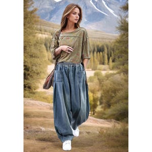 May include: A woman in a long-sleeved olive green top with a floral design and blue denim trousers. The trousers have a wide, loose fit with pockets. She wears white trainers and carries a brown shoulder bag. The background shows a mountain range and trees.