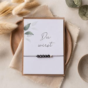 Könnte beinhalten: Eine rechteckige weiße Karte mit dem Text Du wirst in schwarzer Schreibschrift und ein schwarzes Schnurarmband mit runden schwarzen Perlen, die ONKEL buchstabieren, sowie einem kleinen silbernen Sternanhänger auf braunem Papier.
