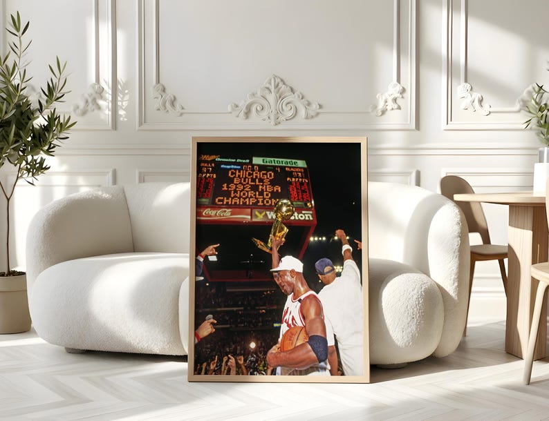 May include: A framed vertical photograph depicting a basketball player in a white jersey holding a gold trophy aloft. The scoreboard in the background displays the text Chicago Bulls 1992 NBA World Champions. The scene captures a stadium celebration.