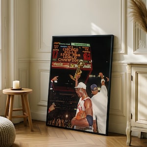 May include: A framed vertical poster featuring a photograph of a basketball player holding a gold trophy aloft. The scoreboard in the background displays the text CHICAGO BULLS 1992 NBA WORLD CHAMPION. The player wears a white jersey and a white cap.