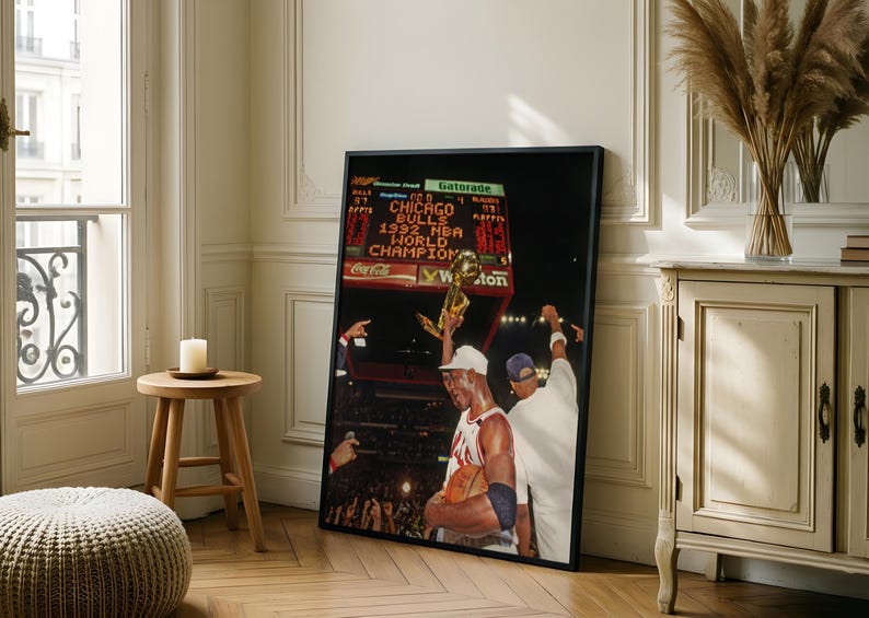 May include: A framed vertical poster featuring a photograph of a basketball player holding a gold trophy aloft. The scoreboard in the background displays the text CHICAGO BULLS 1992 NBA WORLD CHAMPION. The player wears a white jersey and a white cap.