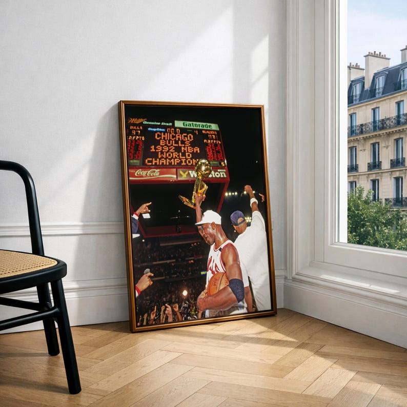 May include: A framed photograph depicting a basketball player in a white jersey holding a gold trophy aloft. The background shows a stadium scoreboard displaying the text CHICAGO BULLS 1992 NBA WORLD CHAMPION, with a crowd visible below the scoreboard.