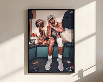 Op de afbeelding: Een ingelijste fotoprint van een basketballer die in een kleedkamer zit, gekleed in een donker shirt, een pet en een kniebrace, terwijl die een gouden kampioenschapstrofee met beide handen vasthoudt en het hoofd ertegenaan laat rusten.