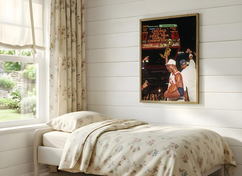 May include: A framed wall art print featuring a photograph of a basketball player holding a trophy with the text Chicago Bulls 1992 NBA World Champion visible on a scoreboard in the background, displayed in a room with floral bedding.