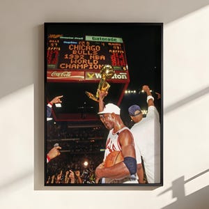 May include: A framed wall art print depicting a basketball player in a white jersey holding a gold trophy aloft. The background shows a stadium scoreboard with the text CHICAGO BULLS 1992 NBA WORLD CHAMPIONS illuminated in bright orange letters.