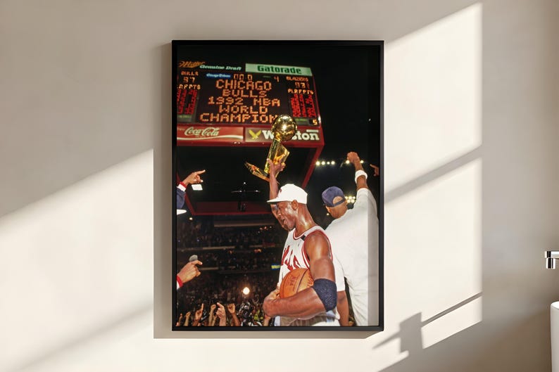 May include: A framed wall art print depicting a basketball player in a white jersey holding a gold trophy aloft. The background shows a stadium scoreboard with the text CHICAGO BULLS 1992 NBA WORLD CHAMPIONS illuminated in bright orange letters.