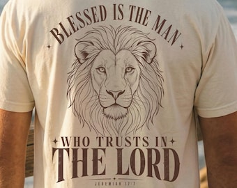 May include: A cream-coloured short-sleeve t-shirt featuring a small dark brown cross on the front left chest. The back displays a large illustration of a lion's head with the text BLESSED IS THE MAN WHO TRUSTS IN THE LORD and JEREMIAH 17:7.