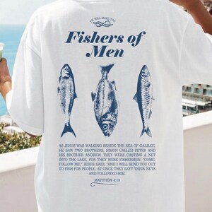 May include: White t-shirt with a blue graphic of three fish and the text "Fishers of Men." The back of the shirt also features a Bible verse. The shirt is a relaxed fit.