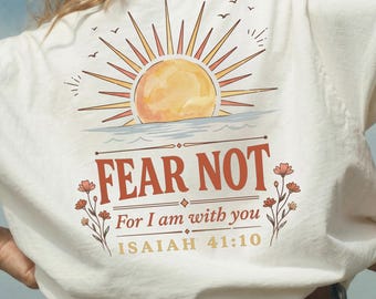 May include: White t-shirt with a sunset graphic and the text "FEAR NOT" along with the Bible verse Isaiah 41:10. The design incorporates a sun, water, and floral elements. The shirt is a relaxed fit.